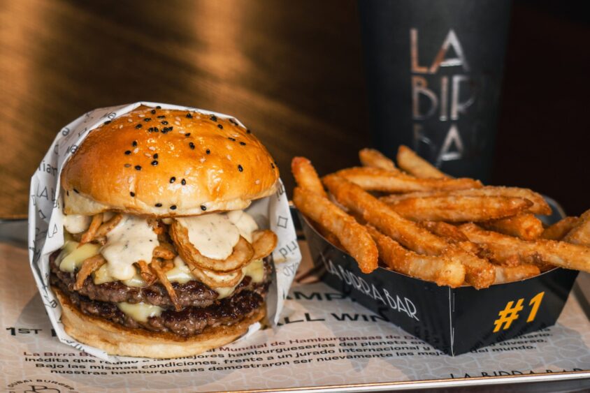 A cheeseburger topped with crispy onions and sauce is served in a sesame bun alongside a tray of golden French fries—an April Flavor Files highlight. A black cup with "La Birra Bar" text is in the background. Lifestyle