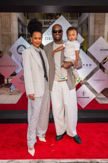 A stylishly dressed woman, man, and young child pose smiling on a red carpet at a Driven By Design event, with a geometric patterned backdrop behind them. Lifestyle