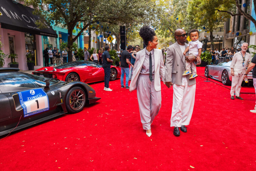 A stylish family, Driven By Design, walks hand-in-hand on a red carpet at a car show, luxury sports cars gleaming behind them as crowds gather among trees and city buildings. Lifestyle