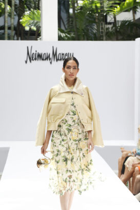 A model walks a runway in a lemon-printed dress with a light yellow jacket draped over her shoulders, holding a metallic gold handbag—a fresh nod to fashion trends. The background displays the Neiman Marcus logo and features lush green plants. Lifestyle
