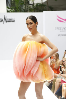 A model walks the runway wearing a voluminous, strapless dress in soft pink and yellow hues, resembling delicate flower petals, as Fashion takes the lead at a show with audience members in the background. Lifestyle