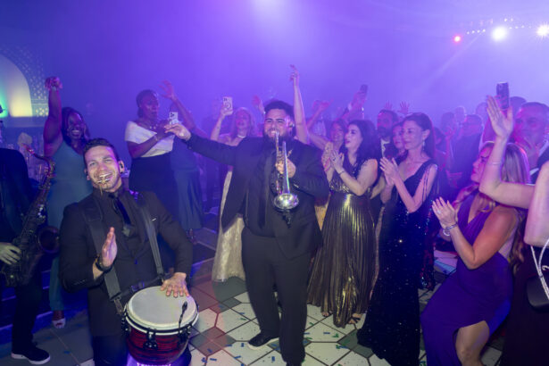 Musicians from Golden Angels play drums and trumpet in the center of a lively crowd in formal attire. People clap, cheer, and take photos under purple stage lights, celebrating a night of real impact. Lifestyle