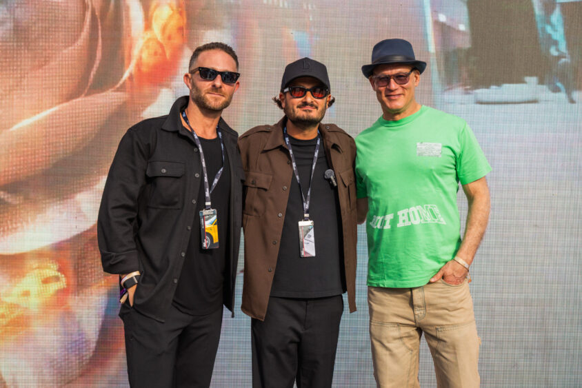 Three men pose together in front of a colorful, abstract background at a Driven By Design event. Two wear dark jackets and sunglasses, while the third sports a green T-shirt, hat, and glasses. All have event badges around their necks. Lifestyle