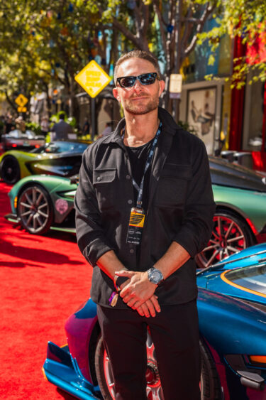 A man wearing sunglasses and a black outfit stands on a red carpet in front of sports cars at an outdoor event, embodying the spirit of Driven By Design. Trees, signs, and buildings are visible in the background. Lifestyle