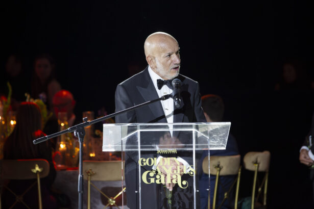 A man in a black tuxedo speaks at a clear podium with “Golden Angel Gala” written on it, celebrating the Real Impact of the Golden Angels as guests sit at decorated tables in the dimly lit event. Lifestyle