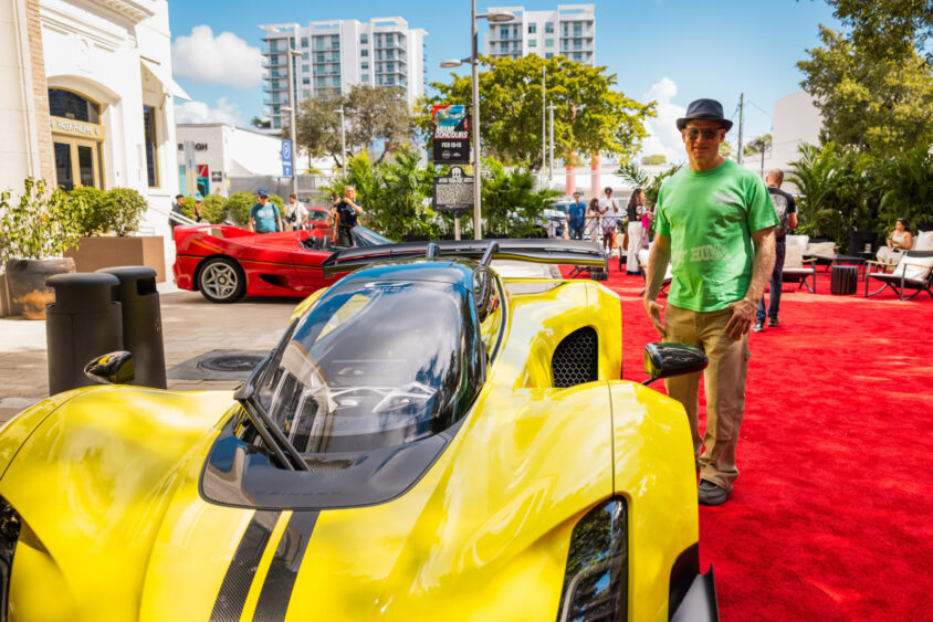 A man in a green shirt and hat stands on a red carpet next to a bright yellow sports car at an outdoor Driven By Design event, with another red sports car, people, trees, and city buildings in the background. Lifestyle