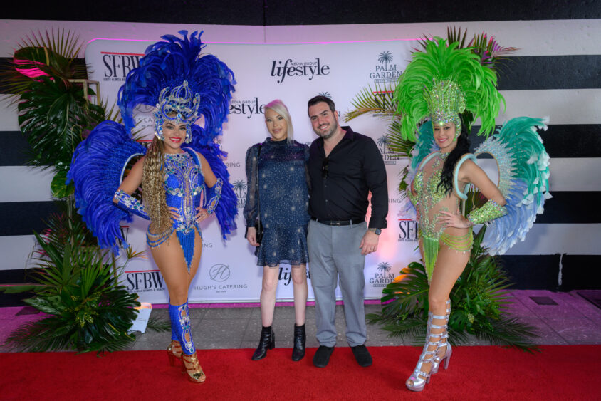Two women in elaborate feathered costumes flank a smiling couple on the red carpet at the March Cover Party, set before a "lifestyle" backdrop and surrounded by palm plants at Palm Garden Event House. Lifestyle