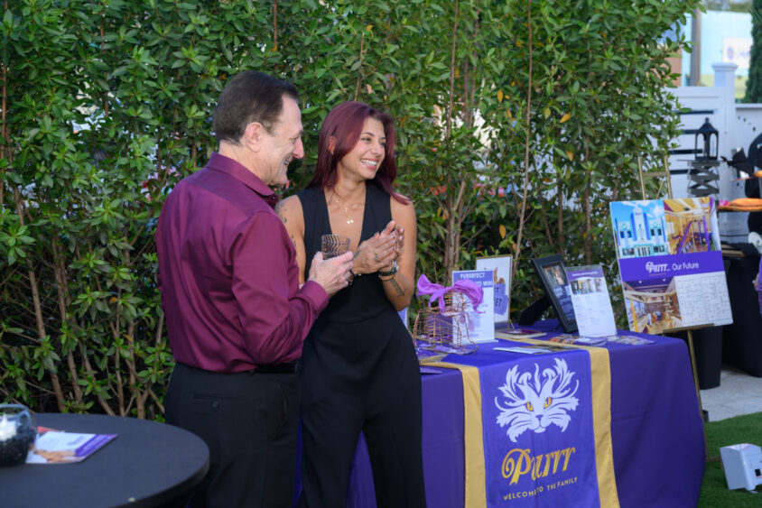 A man and a woman stand smiling and talking beside a table covered with a purple and gold cloth, promotional materials, and brochure stands at the March Cover Party at Palm Garden Event House. They are outdoors, with greenery in the background. Lifestyle