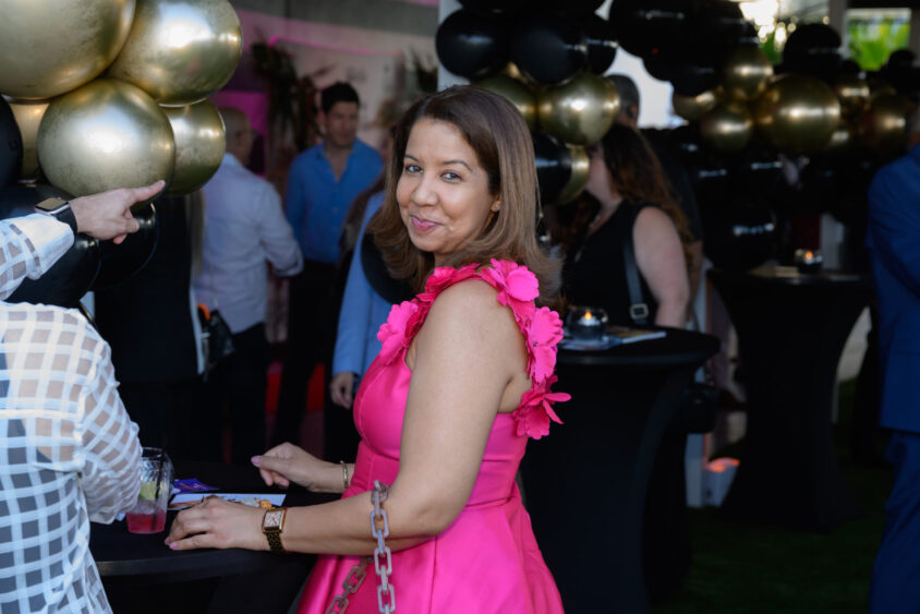 A woman in a bright pink dress with flower details stands and smiles at the camera during the March Cover Party at Palm Garden Event House, with balloons and people celebrating in the background. Lifestyle