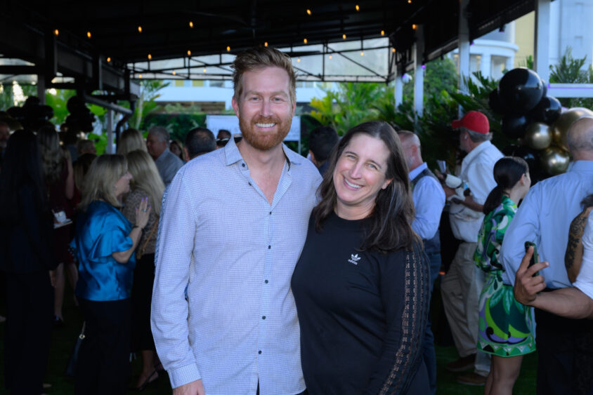 A man and woman stand smiling together at the Palm Garden Event House's March Cover Party, with many people socializing in the background under a covered patio decorated with string lights. Lifestyle