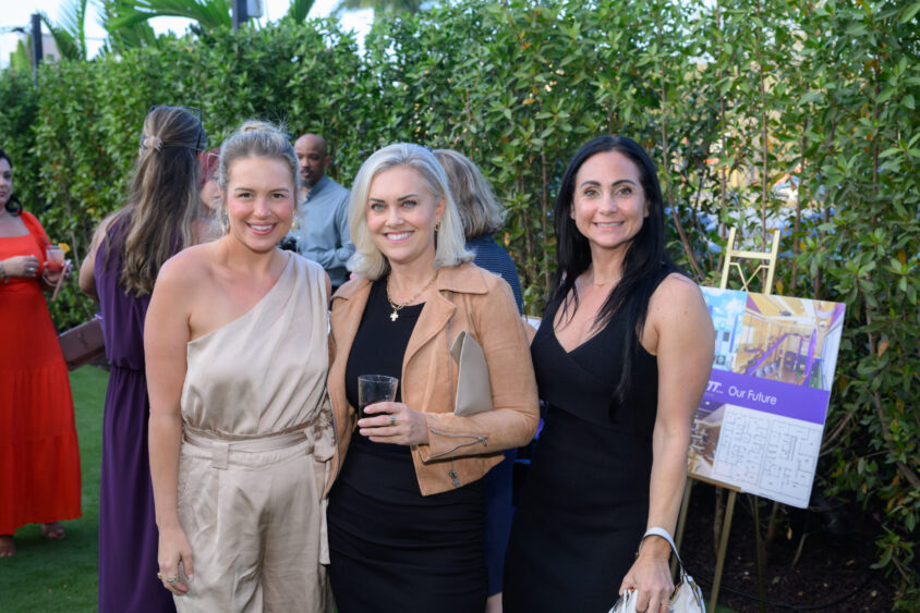 Three women pose and smile at the March Cover Party, held outdoors at Palm Garden Event House. Two hold drinks, while guests socialize on the grass with greenery behind. An easel with a display board stands on the right. Lifestyle