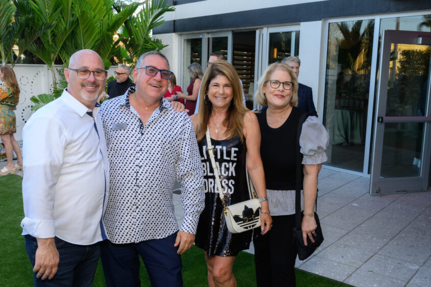 Four adults, three men and one woman, smile outdoors at the Palm Garden Event House during the March Cover Party. They stand on green grass near a building with large windows and lush plants in the background. Lifestyle