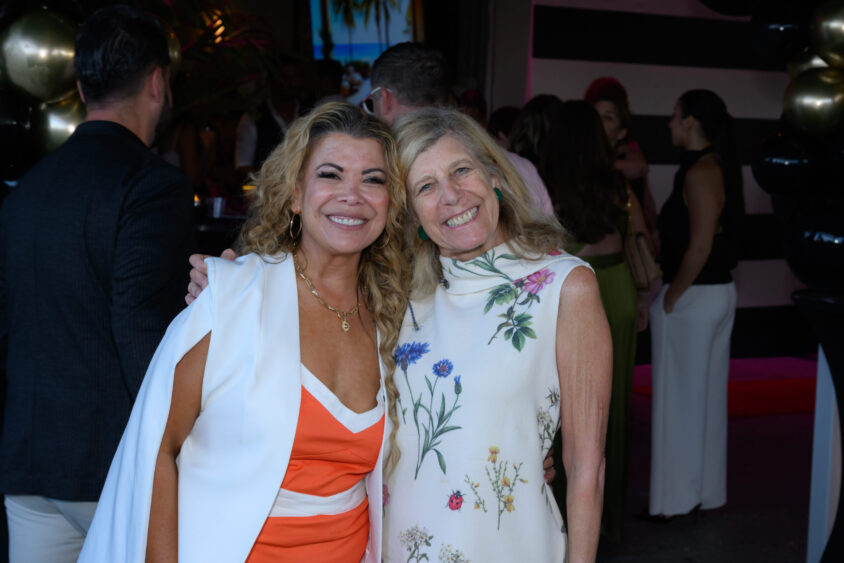 Two women smiling and posing together at the Palm Garden Event House during the March Cover Party. One wears an orange and white dress, the other a sleeveless white floral dress. People and black and gold balloons are in the background. Lifestyle