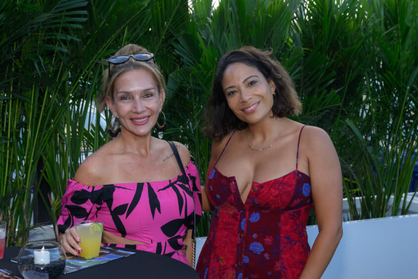Two women stand and smile at the March Cover Party held at Palm Garden Event House. One wears a pink off-shoulder dress with black patterns, the other a red floral dress. Nearby, a table is set with drinks and candles amid lush tropical plants. Lifestyle
