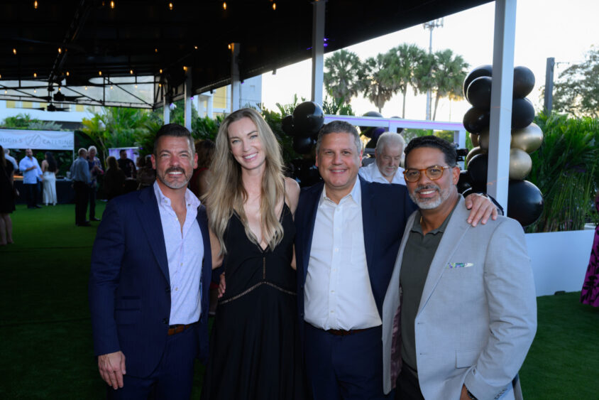 Four people dressed in formal attire smile and pose together at the Palm Garden Event House’s March Cover Party, with green grass, palm trees, festive decorations, and other attendees visible in the background. Lifestyle