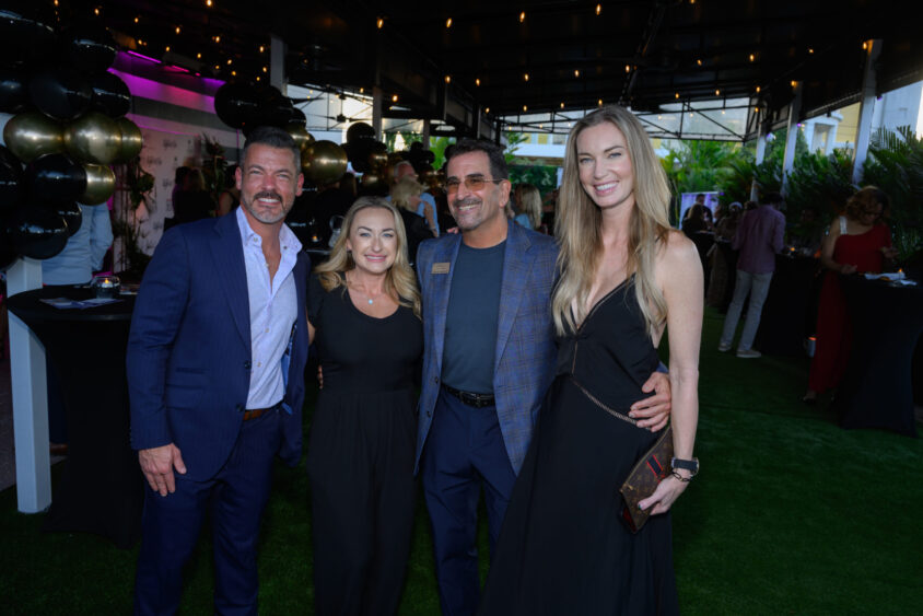 Four people dressed formally smile and pose together at an outdoor March Cover Party at Palm Garden Event House, with string lights, black and gold balloons, and other guests mingling in the background. Lifestyle