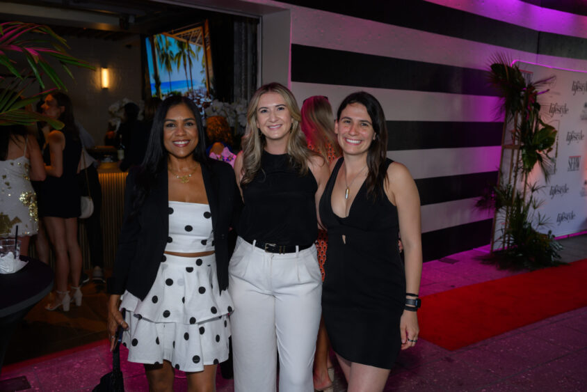 Three women smile and pose together at the March Cover Party with festive lighting at Palm Garden Event House. One wears a polka dot outfit, one a black top and white pants, and one a black dress as other guests mingle in the decorated venue. Lifestyle