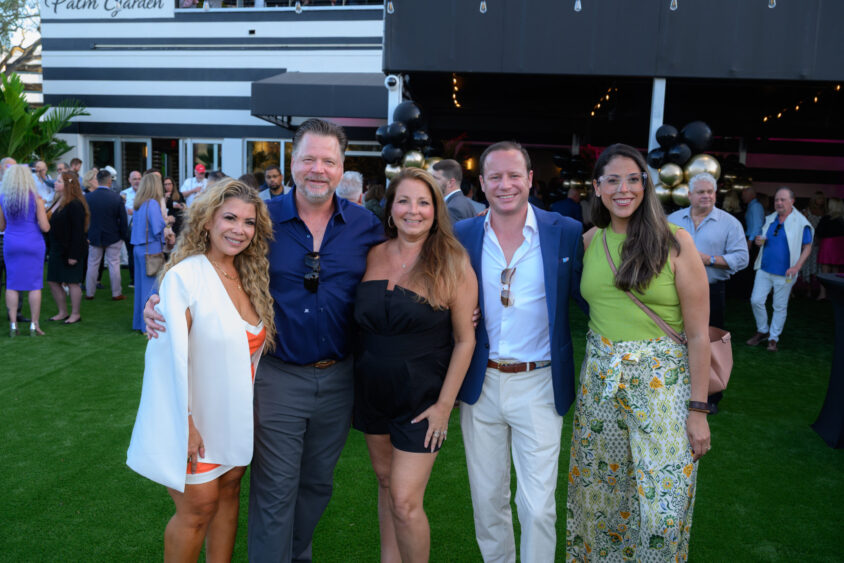 Five people pose and smile together at the Palm Garden Event House’s March Cover Party, with a festive crowd and balloons in the background. Dressed in summer or semi-formal attire, they stand on green grass enjoying the celebration. Lifestyle