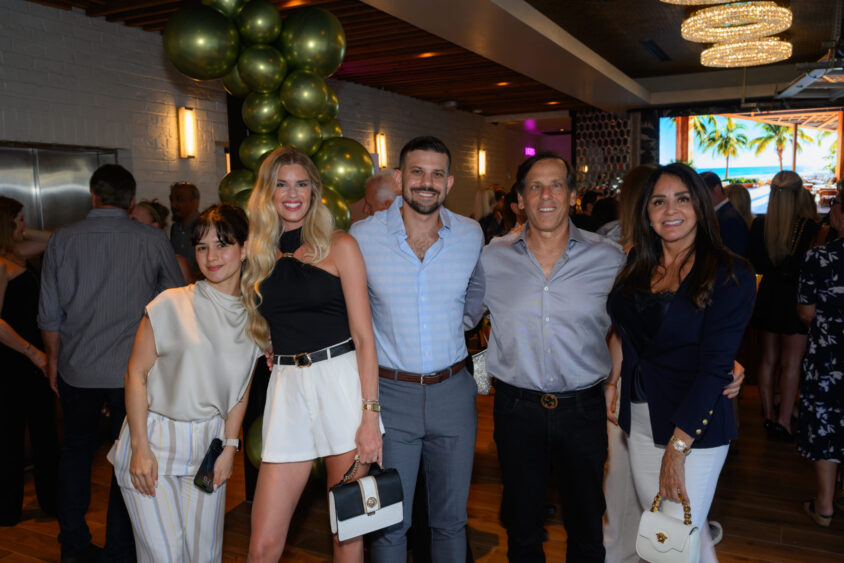 A group of five people stand together smiling at the lively March Cover Party at Palm Garden Event House, with green balloon decorations in the background and casually dressed guests mingling behind them. Lifestyle