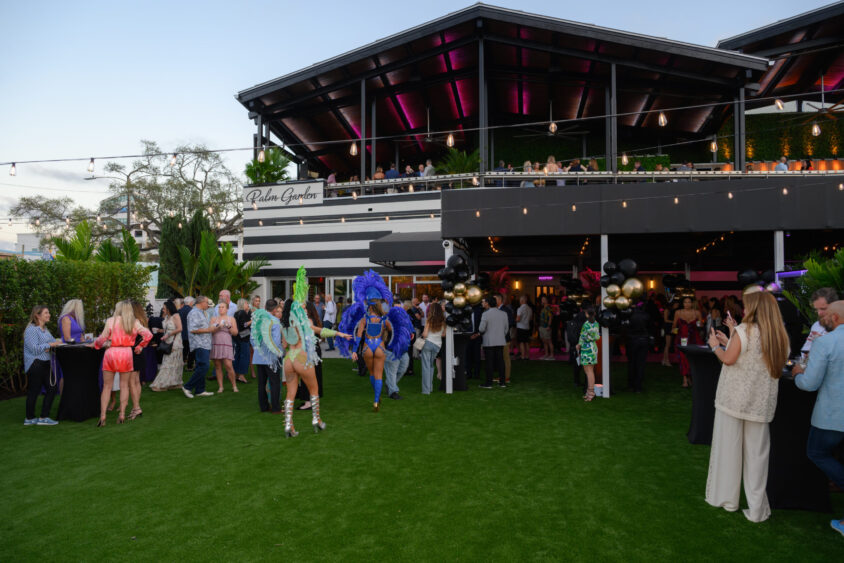 People gather at the March Cover Party on the lawn of Palm Garden Event House, mingling as performers in colorful costumes entertain. The modern, two-level venue glows with lights and balloons while guests socialize and take photos. Lifestyle