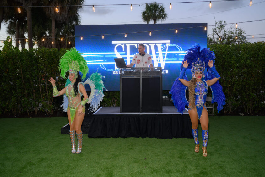 Two dancers in elaborate feathered costumes, one green and one blue, perform on grass at the Palm Garden Event House in front of a DJ booth. Behind them, a lit-up "SERIW" sign shines under string lights at the March Cover Party. Lifestyle