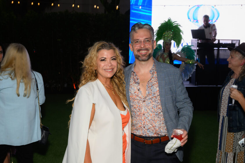 A smiling woman and man pose together at the Palm Garden Event House's March Cover Party. The man holds a drink, while people and a DJ in bright lights mingle in the background amid decorative green costumes. Lifestyle
