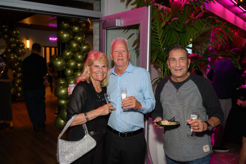 Three smiling adults pose together at the Palm Garden Event House’s March Cover Party, surrounded by festive balloon decorations and pink lighting. The woman holds a sparkling handbag, while the two men hold drinks and a plate of food. Lifestyle