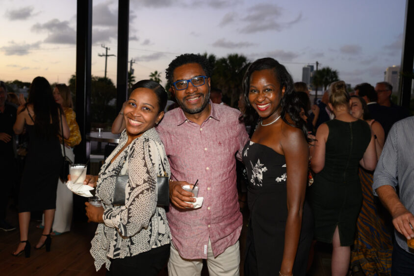 Three smiling people pose closely together at the March Cover Party, held at Palm Garden Event House. The man in the center wears glasses and a pink shirt; the two women beside him hold drinks as a crowd mingles under a dusky sky. Lifestyle