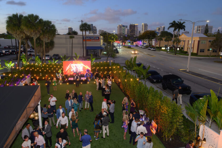 An outdoor evening March Cover Party on a lawn with string lights, a stage with a screen, and groups mingling. Palm trees and city buildings line the busy road in the background at Palm Garden Event House. Lifestyle