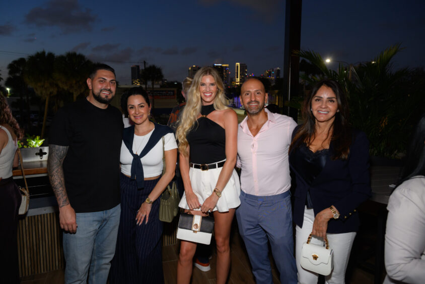 Five adults pose and smile together outdoors in the evening at the Palm Garden Event House, with city lights and palm trees glowing in the background. They are dressed in semi-formal attire, enjoying a relaxed March Cover Party. Lifestyle