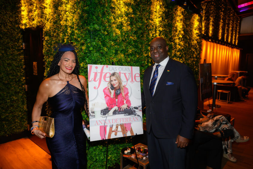 Two people in formal attire stand beside a Las Olas Lifestyle magazine cover featuring a woman DJ, indoors at Palm Garden Event House with lush green wall decor and warm lighting during the March Cover Party. Lifestyle