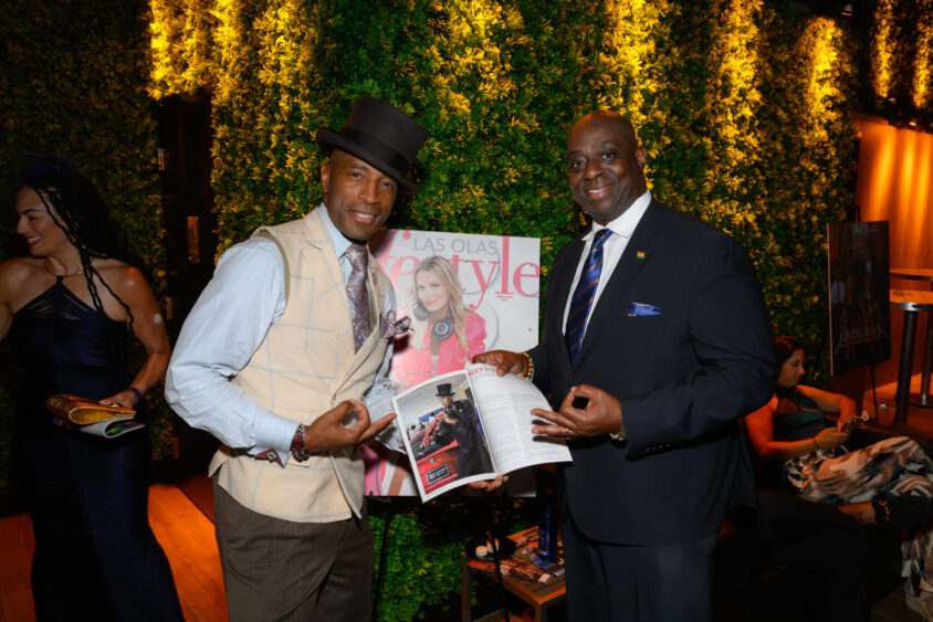 Two men in formal attire smile and pose with a magazine in front of a "Las Olas Lifestyle" cover at the Palm Garden Event House's March Cover Party, while a woman in a blue dress stands against a green foliage wall in the background. Lifestyle