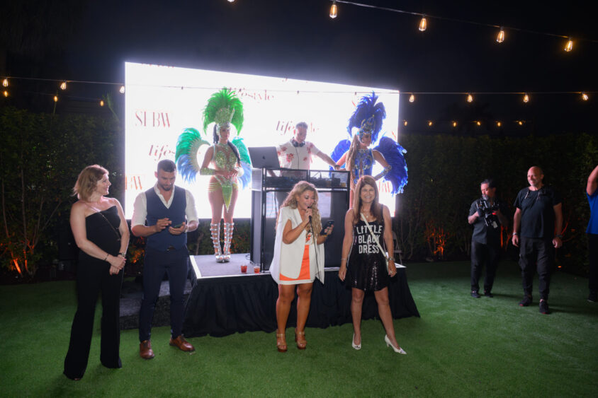 A group of people stand on a lawn at night at the Palm Garden Event House during the March Cover Party, in front of a brightly lit stage where two performers in colorful feather costumes and a DJ entertain. String lights hang overhead as guests chat and take photos. Lifestyle