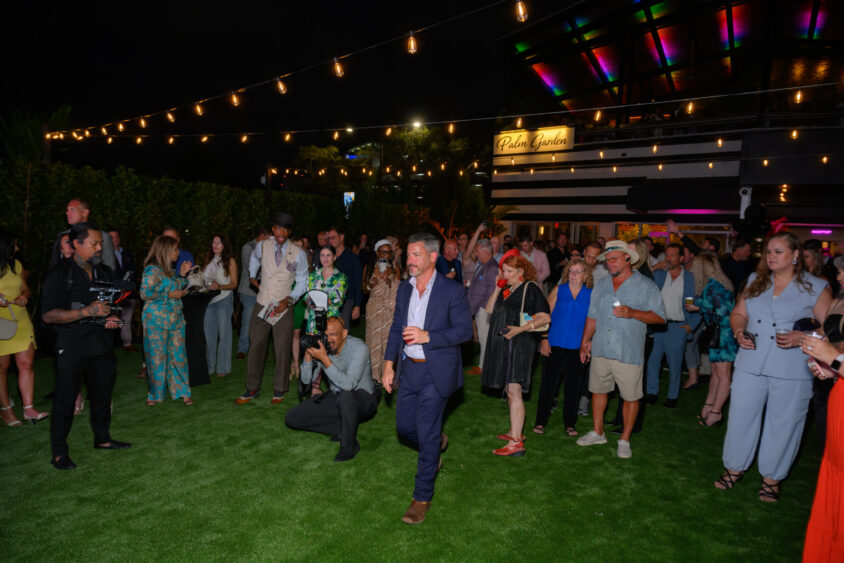 A group of people in semi-formal attire mingle on a grassy lawn at a nighttime March Cover Party, with string lights overhead and the Palm Garden Event House sign glowing in the background. Lifestyle