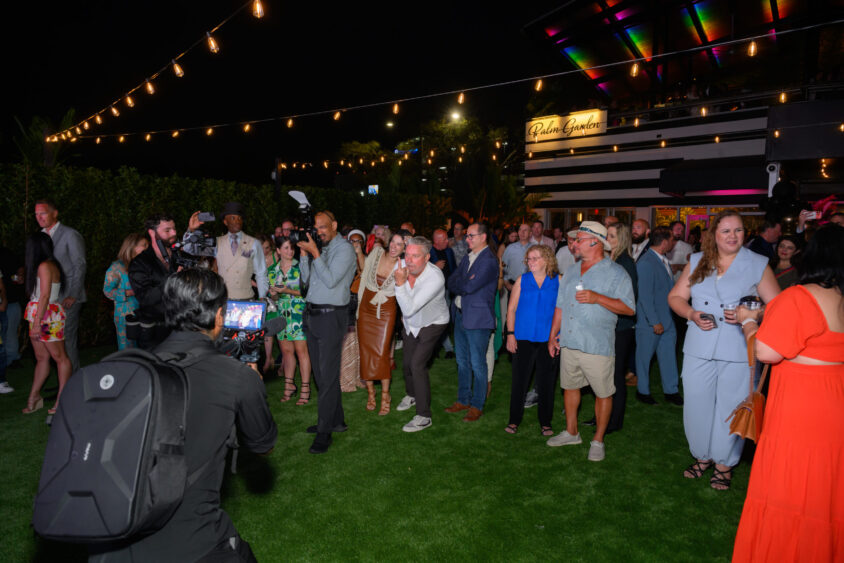A group enjoys an outdoor evening at Palm Garden Event House under string lights, with a photographer and videographer capturing the March Cover Party crowd dressed in semi-formal attire on the grass. Lifestyle
