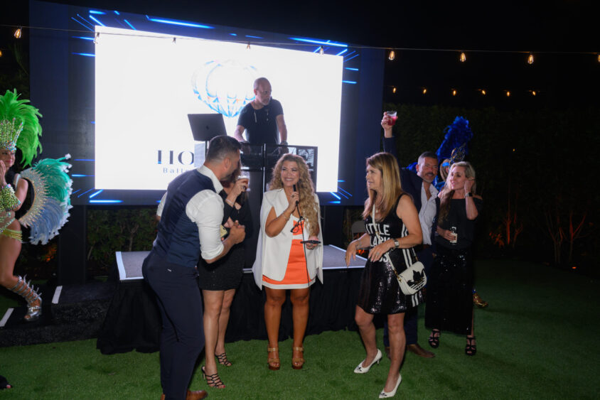A group of people dressed in party attire stand and talk in front of a DJ on an outdoor stage at night at Palm Garden Event House, with bright stage lights and performers in colorful costumes—part of the lively March Cover Party. Lifestyle