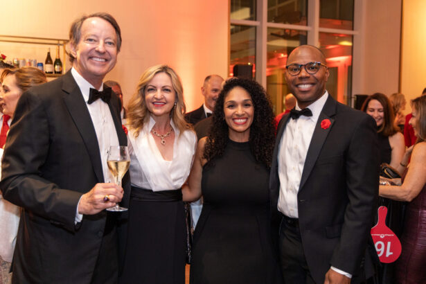 Four people, dressed in formal evening wear, smile and pose together at an indoor philanthropy event. The man on the left holds a glass of champagne; a lively crowd and warm lighting create a welcoming sense of community in the background. Lifestyle