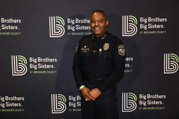 A police officer in uniform stands smiling in front of a backdrop with repeated "Big Brothers Big Sisters of Broward County" logos, ready to make a big impact in the community. Lifestyle