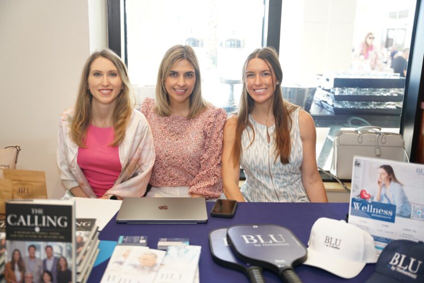 Three women sit at a table with 4KIDS fundraising materials, books, hats, and pickleball paddles branded "BLU." They’re smiling at the Broward Galentine’s event, with a blue cloth on the table and a window display behind them. Lifestyle
