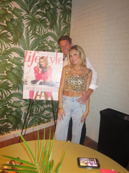 A smiling couple poses indoors in front of a "Las Olas Lifestyle" magazine poster at the March Cover Party. The woman wears a sparkly crop top and jeans, and the man stands behind her, with leafy wallpaper and a white brick wall in the background. Lifestyle