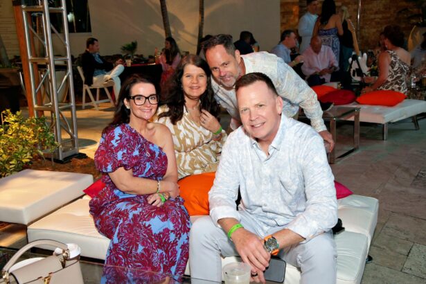 Four adults, two women and two men, sit closely together on white outdoor lounge furniture at a lively, evening social event. The festive, relaxed setting is filled with summer vibes as people in the background chat and enjoy the night. Lifestyle