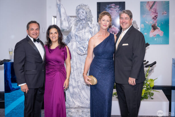 Four adults in formal evening attire pose together, smiling, as Golden Angels making a Real Impact, in front of a marble statue and colorful artwork at an indoor event or gala. Lifestyle