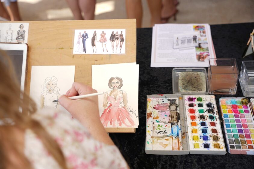 A person paints a fashion illustration of a woman in a pink dress at a table with watercolor paints, sketches, and art supplies during a Broward Galentine’s fundraising event. People stand in the background. Lifestyle