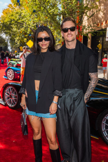 A woman and a man, driven by design, wearing dark sunglasses and black outfits pose together on a red carpet outdoors, with luxury cars and trees visible in the background. Lifestyle