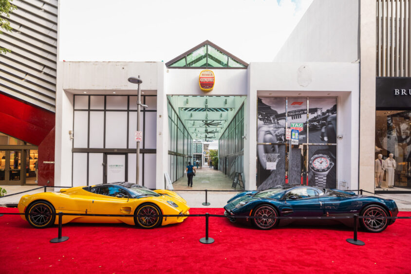 Two luxury sports cars, one yellow and one dark green, are displayed on a red carpet outside a modern building with glass entrance and watch advertisements, showcasing how they are truly Driven By Design. The area is roped off, with a person visible in the background. Lifestyle