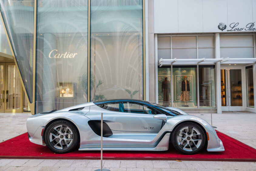 A sleek silver sports car, Driven By Design, is displayed on a red carpet in front of luxury shops like Cartier and Loro Piana in an upscale shopping area. Lifestyle
