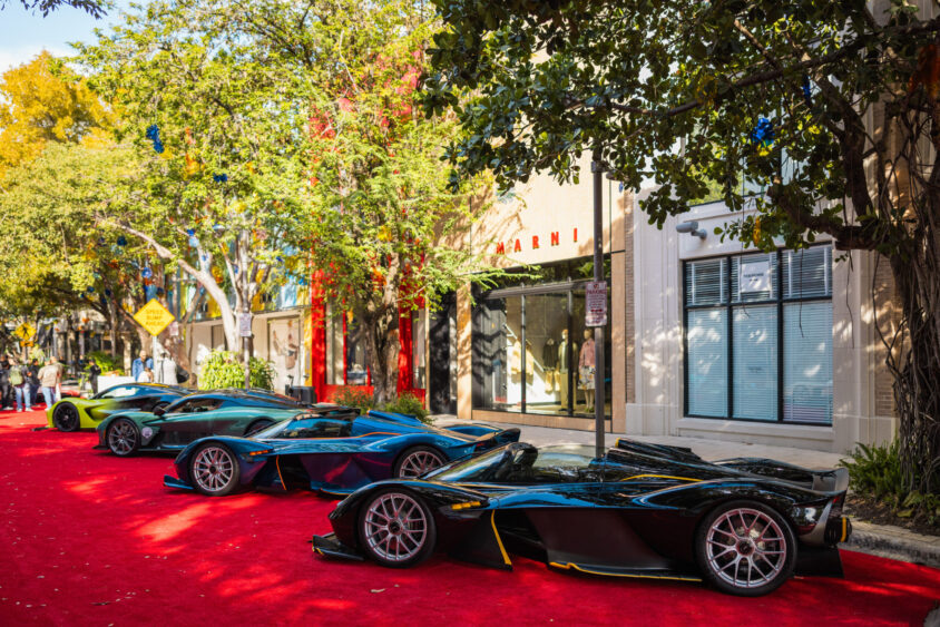 Three sleek, black luxury sports cars—driven by design—are parked side by side on a vibrant red carpet along a tree-lined street, with upscale storefronts and sunlight filtering through the leaves. Lifestyle