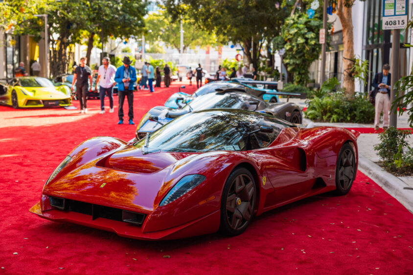 A sleek red sports car, driven by design, is parked on a red carpet outdoors, surrounded by other luxury cars and people. Trees and modern buildings are visible in the background on a sunny day. Lifestyle