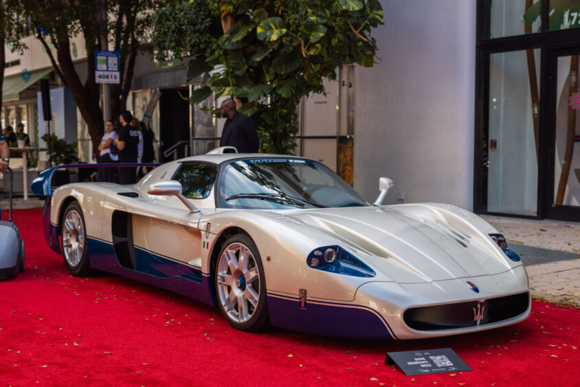 A white and blue Maserati MC12 sports car, driven by design, is displayed on a red carpet outdoors, surrounded by trees and people in the background. Lifestyle
