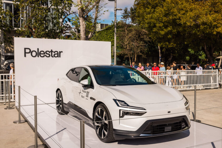 A white Polestar electric car, Driven By Design, is displayed outdoors on a platform with a large Polestar sign behind it. People gather around, while trees and buildings form the backdrop. Lifestyle